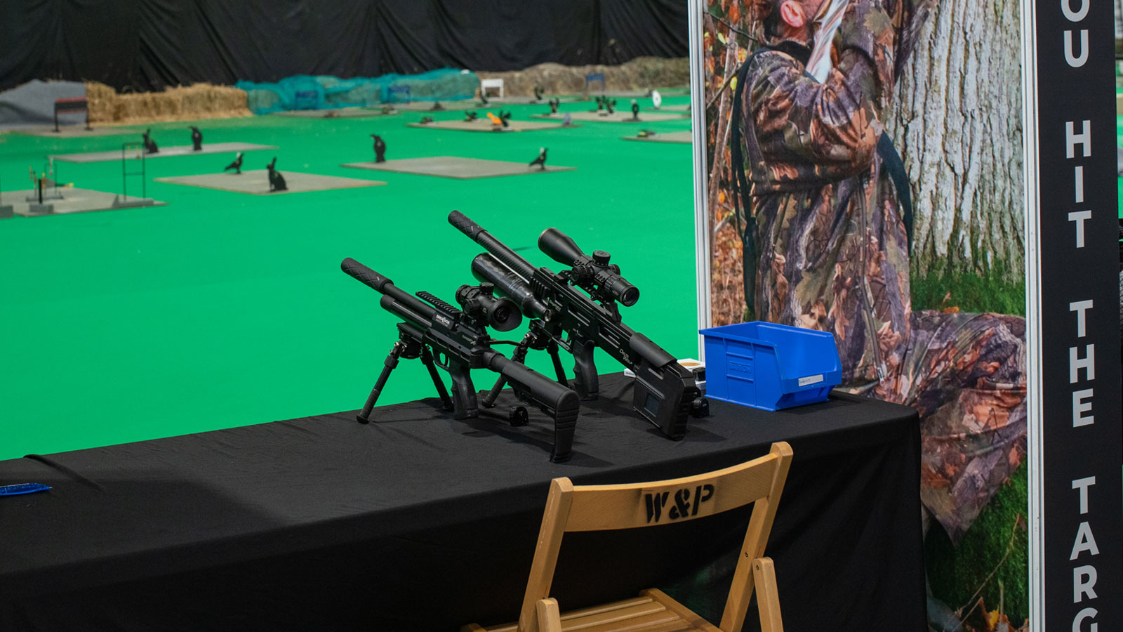Two air rifles sit atop a table infront of an indoor range